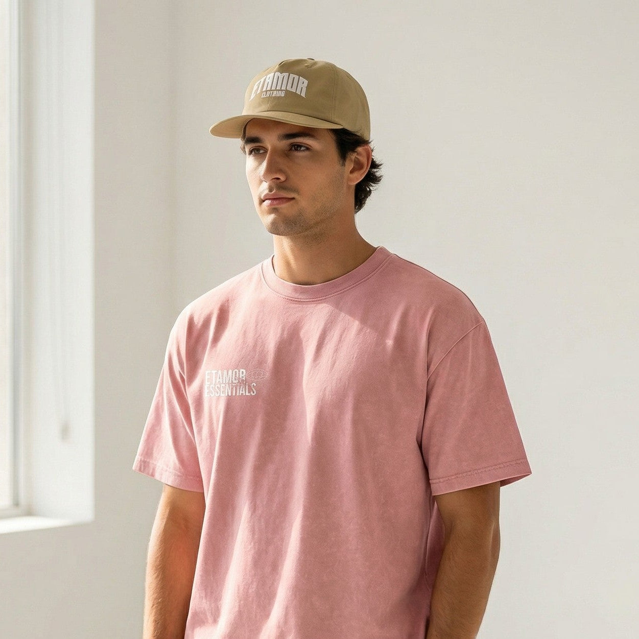 Man wearing a pink t-shirt and gray shorts with a beige cap indoors.