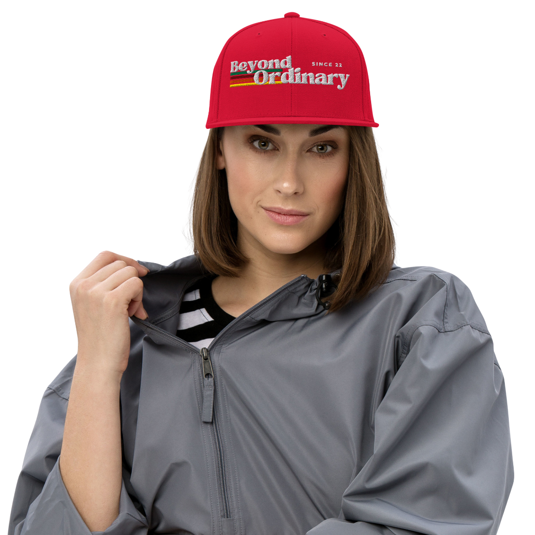 Woman wearing a red 'Beyond Ordinary' cap and gray raincoat on a white background